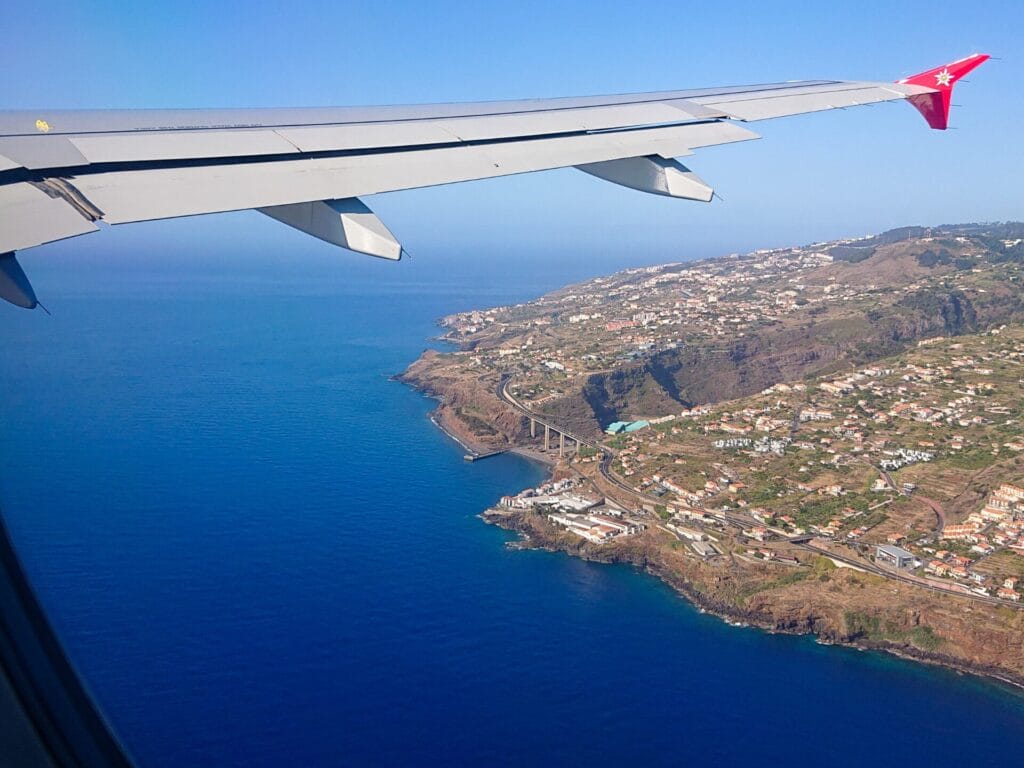 Blick aus dem Flugzeugfenster auf die Küste einer Insel bei wolkenlosem Himmel. Am oberen Bildrand ist der Flugzeugflügel zu sehen